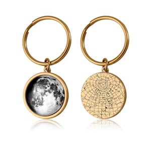 May include: Two gold-colored keychains. One keychain features a round pendant with a black and white image of the moon. The other keychain has a round pendant with a map design. Both pendants are attached to a gold-colored key ring.