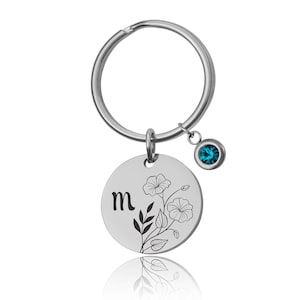 以下が含まれることがあります： 花柄と文字「m」が刻まれた丸いチャームが付いたシルバーカラーのキーホルダー。キーホルダーリングの下には、小さなティール色の宝石がぶら下がっています。花柄のデザインには花と葉が含まれています。