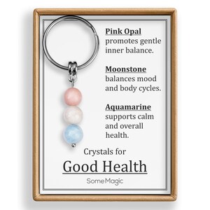 May include: A keychain with three gemstone beads in pink, white, and blue. The card reads "Crystals for Good Health" and lists the benefits of Pink Opal, Moonstone, and Aquamarine. The keychain is in a wooden frame.