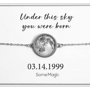 Può includere: Un braccialetto argentato con un ciondolo circolare raffigurante un'immagine dettagliata della luna. Il braccialetto è presentato su una carta bianca con il testo "Under this sky you were born", la data "03.14.1999" e il nome del marchio "SomeMagic".