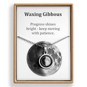 May include: A silver-tone necklace with a pendant featuring a moon phase design, presented in a framed display. The frame contains the text "Waxing Gibbous" and a motivational quote. The background shows a grayscale image of the moon.