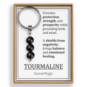 May include: A black tourmaline gemstone keychain with three round beads. The keychain is attached to a card with text that reads "Provides protection, strength, and prosperity..." and "TOURMALINE SomeMagic."