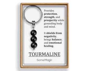 May include: A black tourmaline gemstone keychain with three round beads. The keychain is attached to a card with text that reads "Provides protection, strength, and prosperity..." and "TOURMALINE SomeMagic."