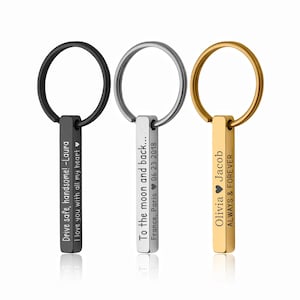 May include: Three personalized keychains with engraved text. One is black, one is silver, and one is gold. The black keychain reads "Drive safe, handsome! -Laura I love you with all my heart." The silver keychain reads "To the moon and back... France, Paris 06 23 2018." The gold keychain reads "Olivia Jacob ALWAYS & FOREVER..."