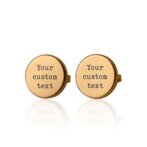 Peut inclure: Une paire de boucles d'oreilles rondes dorées. Chaque boucle d'oreille porte l'inscription "Your custom text" gravée sur le devant. Les boucles d'oreilles sont présentées sur fond blanc avec un léger reflet.