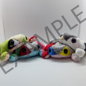 Puede incluir: Tres juguetes de peluche con forma de rana. La primera rana es verde con detalles rosas y tiene ojos negros. La segunda rana es azul, roja y gris con ojos negros. La tercera rana es rosa, gris y amarilla con ojos negros.