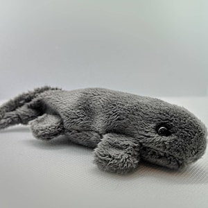 May include: A grey plush toy shaped like a whale with a single black eye. The whale is lying on its side with its tail curled up.