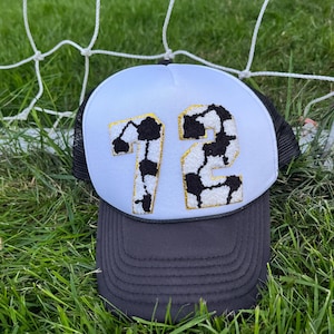 May include: A trucker hat with a white front panel and a black mesh back. The front features the number "72" in a black and white cow print design with gold trim. The brim is dark gray, and the hat is set against a backdrop of green grass and a soccer net.