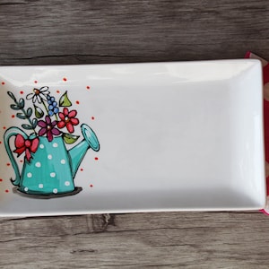 Peut inclure: Un plat de service rectangulaire en céramique blanche avec un motif peint à la main d'un arrosoir bleu à pois blancs rempli de fleurs colorées. Le plat est sur une surface en bois.