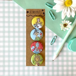May include: Four round pin badges with illustrations of characters from The Wizard of Oz. The badges are attached to a brown card with the word "AIMANTS" at the top. The background is a light green and white checkered pattern.