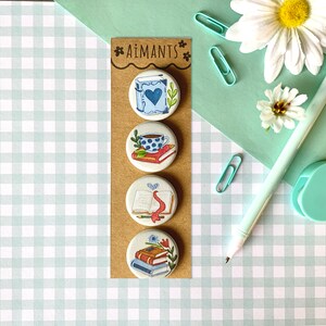 May include: Four round, illustrated buttons on a brown card. The buttons feature book-themed designs, including a book with a heart, a cup of coffee on books, an open book with a ribbon, and a stack of books. The card has the text "AIMANTS" at the top.
