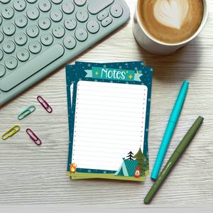May include: A blue and green notepad with a camping theme. The notepad has a white lined interior and the text "Notes" in a white banner with stars. The notepad is on a light wood surface with a keyboard, a cup of coffee, and two pens.