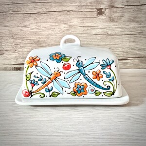Peut inclure: Un beurrier en céramique blanche avec un couvercle orné d'un motif peint à la main de libellules bleues et orange avec des fleurs dans des tons d'orange, jaune, bleu et vert.