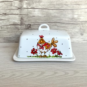 May include: White ceramic butter dish with a lid featuring a hand-painted design of a brown and red chicken with black polka dots and red flowers.