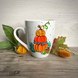 以下が含まれることがあります： 白いセラミック製のマグカップで、手描き風の3段に重なったオレンジ色のカボチャのイラストが描かれています。グレーの水玉模様のリボン、緑色の蔓、小さなヒマワリが添えられています。マグカップには白い持ち手があり、表面全体に小さな黒いドット模様が散りばめられています。