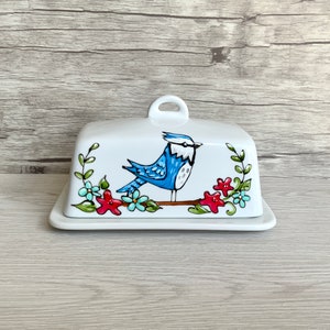 May include: White ceramic butter dish featuring a bluebird and floral design. The bird is perched on a branch with red and blue flowers.