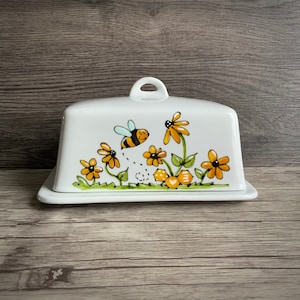 May include: White ceramic butter dish featuring a bee and yellow flowers design. The bee is flying and has black stripes. The flowers are yellow with brown centres.