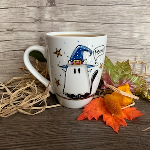 Puede incluir: Una taza de cerámica blanca con una ilustración pintada a mano de un gato disfrazado de fantasma, con un sombrero de mago azul con una hebilla amarilla. El gato está rodeado de estrellas amarillas y el texto Meooo! aparece en una burbuja.