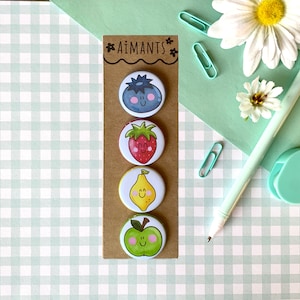 Peut inclure: Un ensemble de quatre badges ronds sur le thème des fruits. Les badges représentent une myrtille, une fraise, un citron et une pomme, chacun avec un visage souriant. Le nom de la marque "Aimants" est imprimé en haut de la carte.