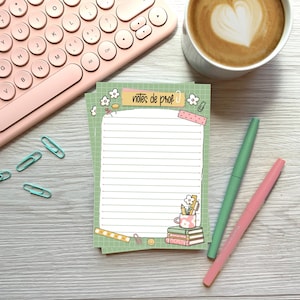 May include: A notepad with the text "notes de prof" in a whimsical design, featuring a grid background, flowers, and a stack of books. A pink keyboard, pens, and a cup of coffee with latte art are also visible.