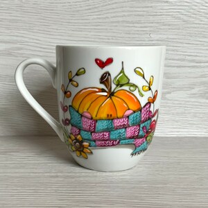 Peut inclure: Un mug en céramique blanche orné d'une illustration peinte à la main représentant une citrouille orange enveloppée dans une écharpe en tricot bleue et rose. Le motif inclut un petit cœur rouge, des feuilles vertes et des fleurs jaunes.