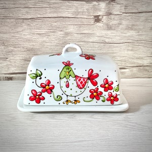 May include: White ceramic butter dish with a lid featuring a hand-painted green and red chicken with red flowers and green leaves.