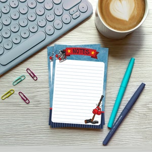 May include: A blue and white notepad with a hockey theme. The notepad has a red ribbon with the word "NOTES" printed on it. There are hockey skates, a hockey stick, and a hockey glove on the notepad. The notepad is on a wooden surface with a cup of coffee, a keyboard, and paper clips.