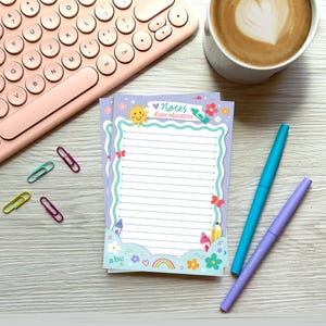 Peut inclure: Bloc-notes avec le texte "Notes d'une éducatrice", bordé de fleurs, papillons et arc-en-ciel. Il y a aussi un clavier rose, du café, des stylos et des trombones.