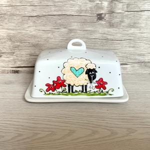 May include: A white ceramic butter dish with a lid. The dish is decorated with a sheep design, featuring a cream-coloured body and a turquoise heart. Red star-shaped flowers and black polka dots add to the whimsical design. Includes a handle.