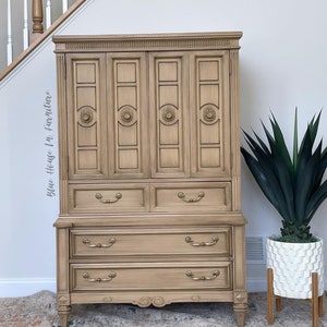 May include: A vintage wooden cabinet with a light brown finish. The cabinet has four doors with brass hardware and four drawers with brass pulls. The cabinet is standing on a rug with a floral pattern.
