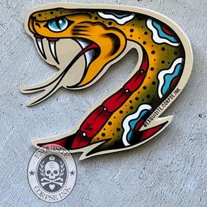 May include: A die-cut sticker featuring a stylised snake head with yellow skin, black spots, and a bright blue eye. The snake has an open mouth with white fangs and a red underbelly. The text EXQUISITE.CORPSE.INK is printed on the sticker and a logo.