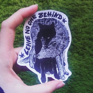May include: A black and white sticker with a wolf illustration. The sticker says "Leave no one behind" with a star on either side of the text.