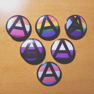 Puede incluir: Seis botones redondos con un símbolo de anarquía en el centro. Cada botón tiene un esquema de color diferente, incluyendo rosa, morado, azul, verde, amarillo y negro.