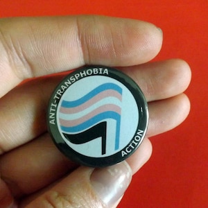 Puede incluir: Un botón blanco y negro con el texto "Anti-Transfobia Action" y una bandera del orgullo transgénero estilizada.