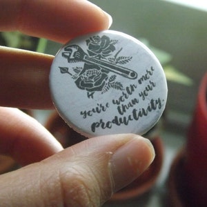 Puede incluir: Un botón redondo blanco con una ilustración negra de una llave inglesa y rosas. El botón tiene el texto "you're worth more than your productivity" en un estilo manuscrito. El botón está sostenido entre los dedos.