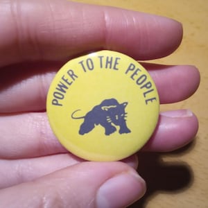 Può includere: Un bottone giallo con una silhouette di pantera nera e il testo "POWER TO THE PEOPLE" in nero.