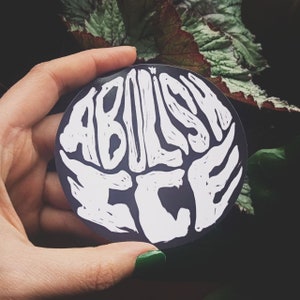 Può includere: Un adesivo bianco e nero con le parole "Abolish ICE" scritte in un carattere stilizzato.