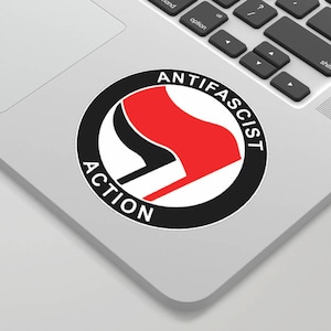 May include: Black and white circle with the text "Anti-Fascist Action" around the edge. Inside the circle is a red and black symbol with a red fist holding a flag.