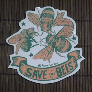May include: A white sticker with three green and pink bees and a pink banner that says "Save the Bees" with the text "@praxisstudio" below the banner.