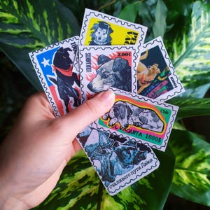 Puede incluir: Una mano sosteniendo una colección de sellos postales con temática de perros. Los sellos presentan varias razas de perros y diseños, con texto que incluye "LOUKANIKOS", "Chile" y "1000$". Los sellos son coloridos y están sobre un fondo de follaje verde.