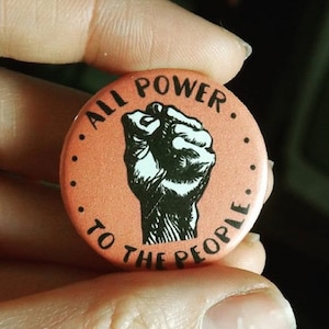 Puede incluir: Un botón de color melocotón con una ilustración en blanco y negro de un puño en alto. El texto "All Power To The People" está escrito en un círculo alrededor del puño.