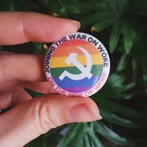 Op de afbeelding: Een ronde knop met een regenboogachtergrond en een wit hamer-en-sikkel symbool. De tekst op de knop luidt "Joining the War on Woke On the Side of Woke."