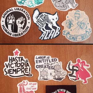 May include: Two sets of stickers with various political and social messages. The first set includes stickers with messages like "Adopt Don't Shop", "Animal Liberation, Human Liberation", "All Cats Are Beautiful", "Eat the Oppressor", and "Save the Bees". The second set includes stickers with messages like "Hasta la Victoria Siempre", "Labor is Entitled to All it Creates", "No War but Class War", and "Solidarity Between Workers Always!"