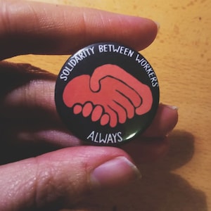 Peut inclure: Un bouton noir avec un graphique de poignée de main rouge et le texte "Solidarité entre les travailleurs toujours".