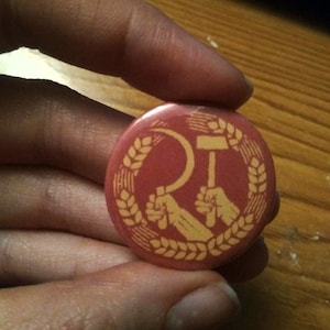 Puede incluir: Un botón redondo rojo con un símbolo amarillo de martillo y hoz, rodeado por una corona de trigo. El diseño incluye dos manos sosteniendo el martillo y la hoz. El botón se sostiene entre los dedos.