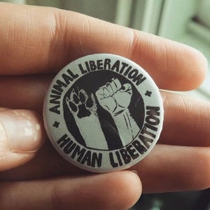 May include: A white button with black text and images. The text reads "Animal Liberation Human Liberation". The images are a raised fist and a paw print.