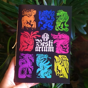 Könnte beinhalten: Ein schwarzes Büchlein mit dem Titel "Bestiarium" mit farbenfrohen Illustrationen von Fabelwesen. Das Cover zeigt verschiedene Kreaturen in leuchtenden Farben, darunter ein Pferd, ein Wolf, ein Löwe und ein Drache. Das Büchlein ist mit einer goldenen Schnur gebunden.