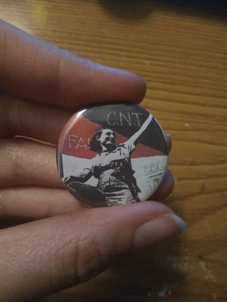 May include: A black and white button with a red and black flag in the background. The flag has the text "CNT" and "FA" visible. A person is raising their fist in the air.