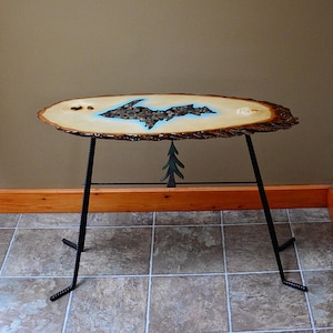 May include: A wooden table with a live edge top featuring a blue and brown inlay of the Upper Peninsula of Michigan. The table has a black metal base with a tree design.