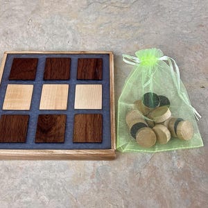 May include: A wooden game board with a gray and blue inlay, featuring a 3x3 grid of squares. The board is made of light and dark wood. A green mesh bag filled with wooden game pieces is next to the board.